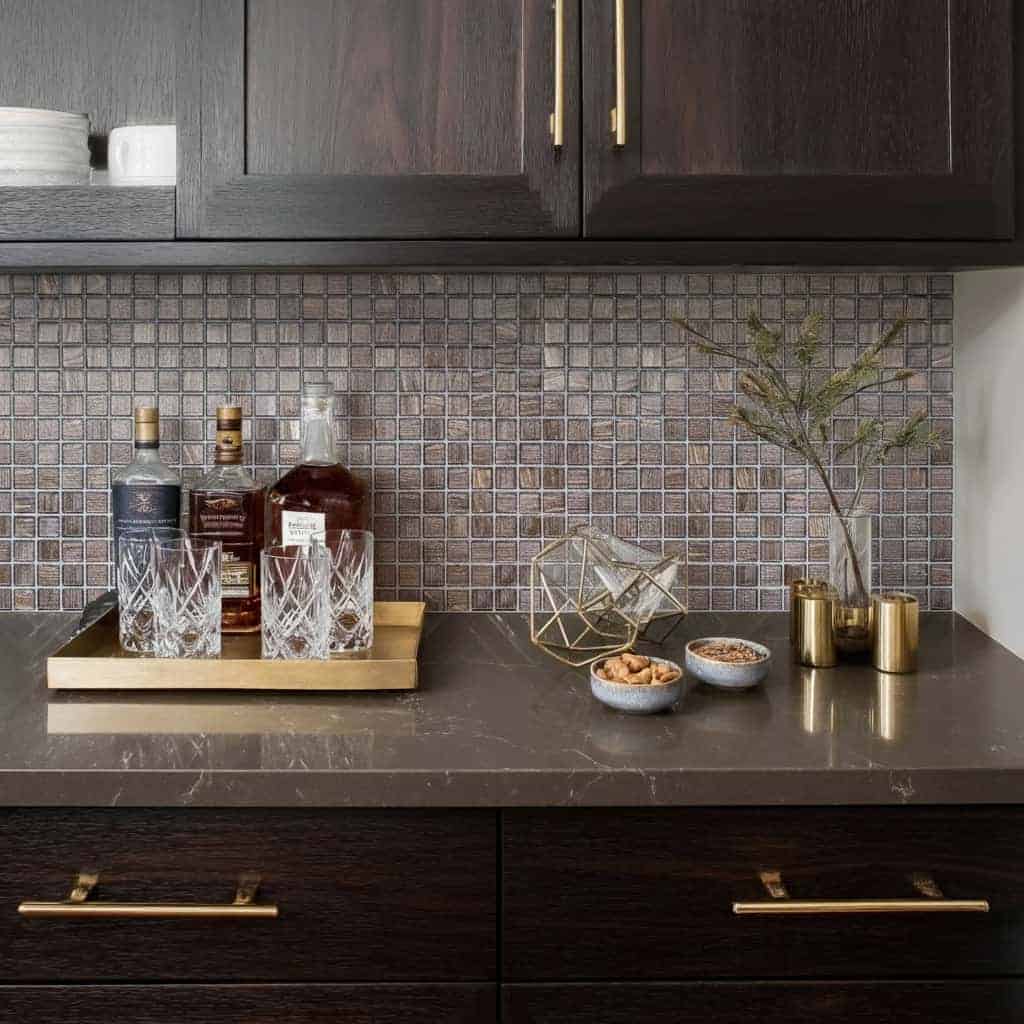 Glass tile backsplash behind a dark kitchen countertop with bottles, bowls, and a plant in gold vases.
