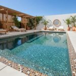 A modern outdoor pool featuring a glass mosaic tile interior, surrounded by wooden and white lounge furniture, with potted pl.