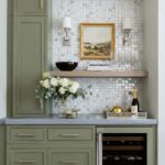 Green kitchen with glass mosaic backsplash, decorative flowers, and wine bottle.