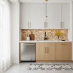 A modern kitchen featuring peel and stick wall tiles, light wood lower cabinets, and white upper cabinets with gold handles.