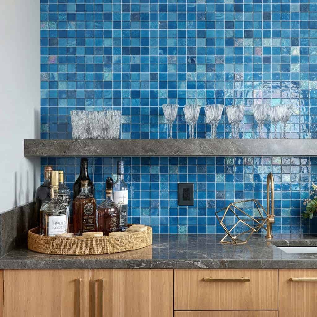 A vibrant blue glass tile backsplash in a contemporary kitchen, paired with a dark countertop, gold faucet, and decorative gl.