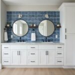 Blue glass tile backsplash with two round mirrors and white cabinetry in a contemporary bathroom.