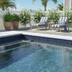 A beautiful pool area featuring glass mosaic tiles, lounge chairs, potted plants, and a white picket fence under a sunny sky.