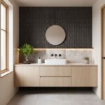 Black textured tile wall behind a white sink with a marble backsplash, light wood vanity, and potted plants.