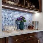Glass tile backsplash with purple flowers in a modern kitchen, featuring a marble countertop and decorative glassware.