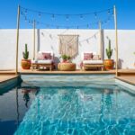 A stylish outdoor seating area featuring glass tile backsplash, pool, and colorful cushions under string lights.