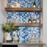 Blue glass tile backsplash with two wooden shelves holding bottles and candles, complemented by decorative vases and glasswar.