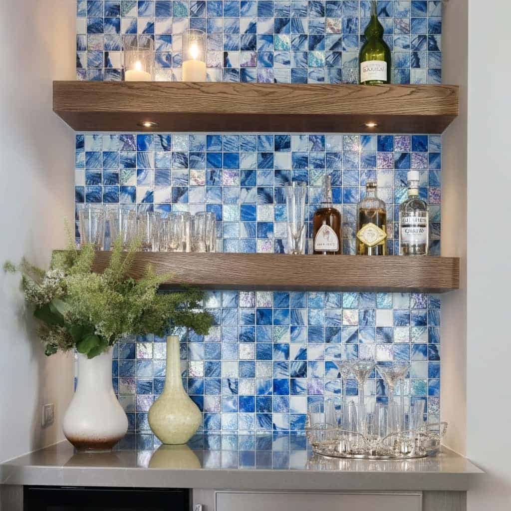 Blue glass tile backsplash with two wooden shelves holding bottles and candles, complemented by decorative vases and glasswar.