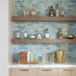 Stylish glass tile backsplash with floating shelves holding bottles and decor, complemented by gold accents and a white cabin.