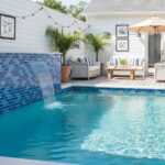 Blue mosaic tile waterfall, textured pool surface, and outdoor furniture with striped cushions.