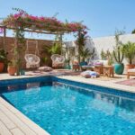 Bright blue mosaic tile pool with surrounding wooden deck and vibrant potted plants.