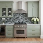 Mint green kitchen cabinets with gold handles, gray mosaic tile backsplash, and stainless steel oven.