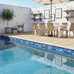 Bright blue mosaic tile border along the pool edge, textured wood decking, and neutral-toned wicker chairs.