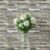 White flowers with green leaves in a clear glass vase, set against a textured gray and beige wood-look tile wall.