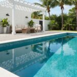 Poolside outdoor seating area with white wooden pergola and lush greenery.